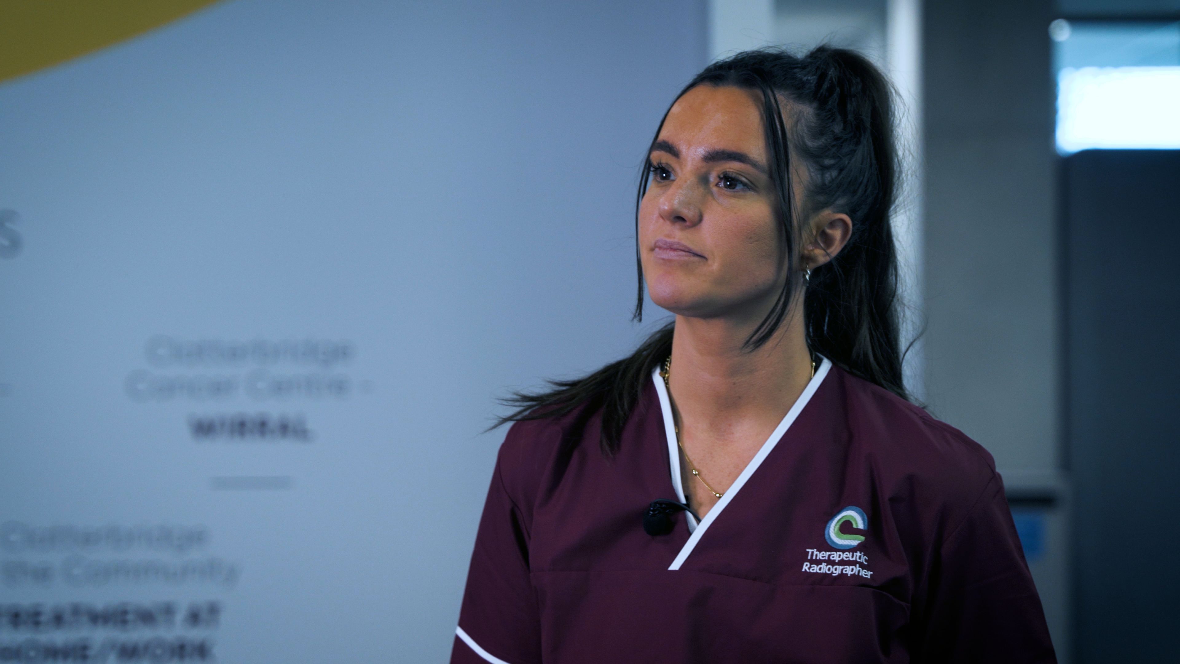Aimee Clark in maroon scrubs with a clip-on microphone, in front of a Clatterbridge Cancer Centre backdrop