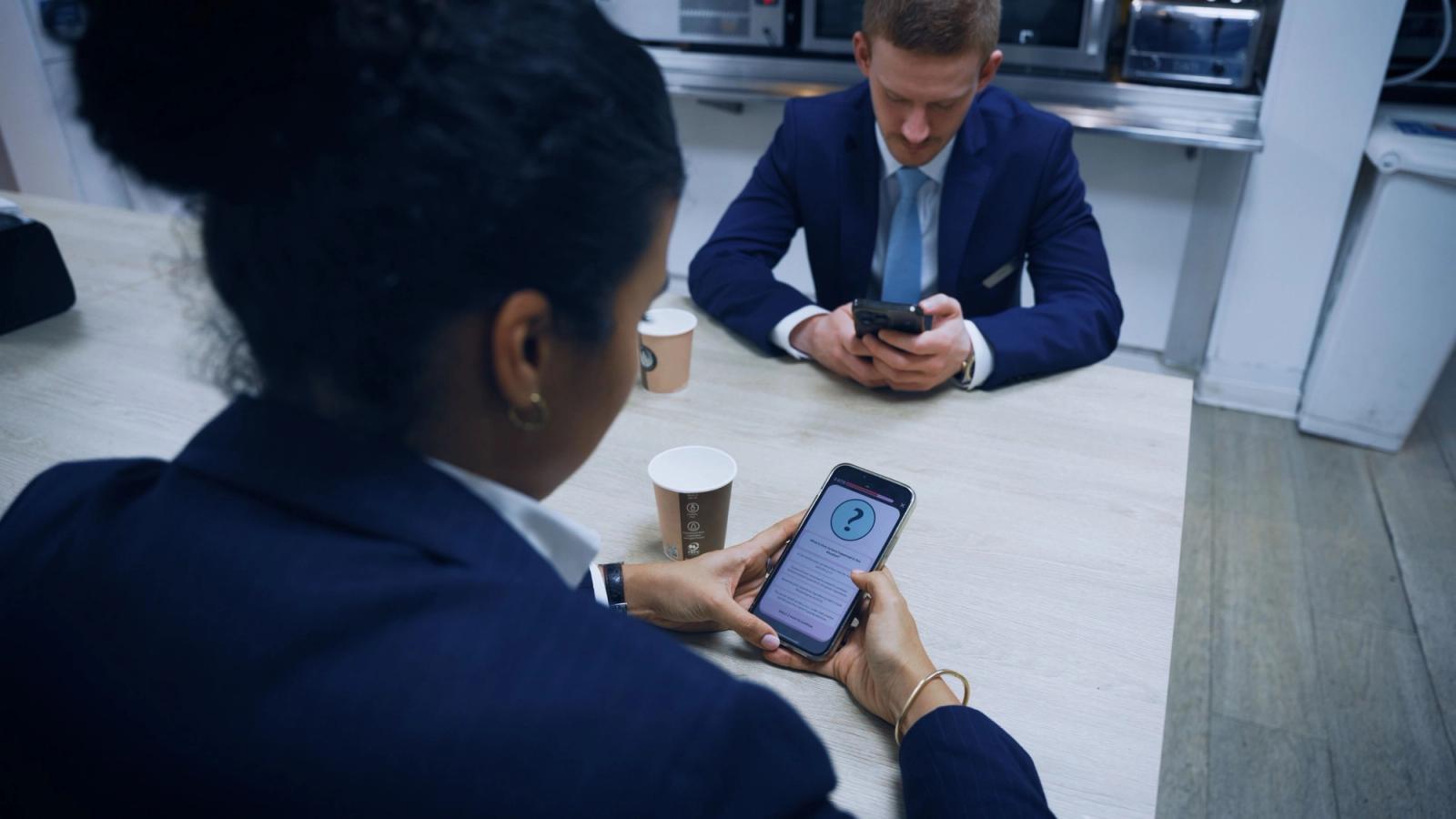 Two people in business suits sit at a table with coffee, both using phones; one screen shows a question-mark icon