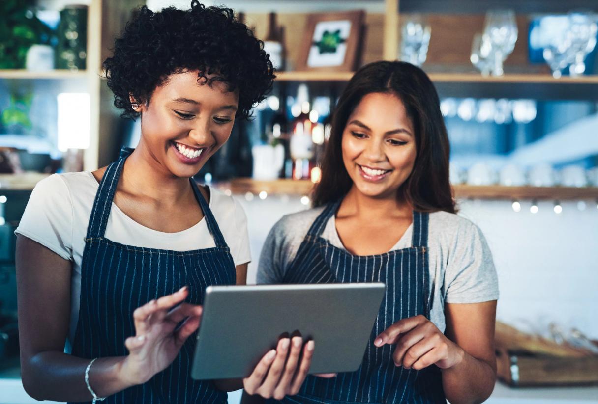 Two smiling café staff in striped aprons looking at a tablet together behind the counter