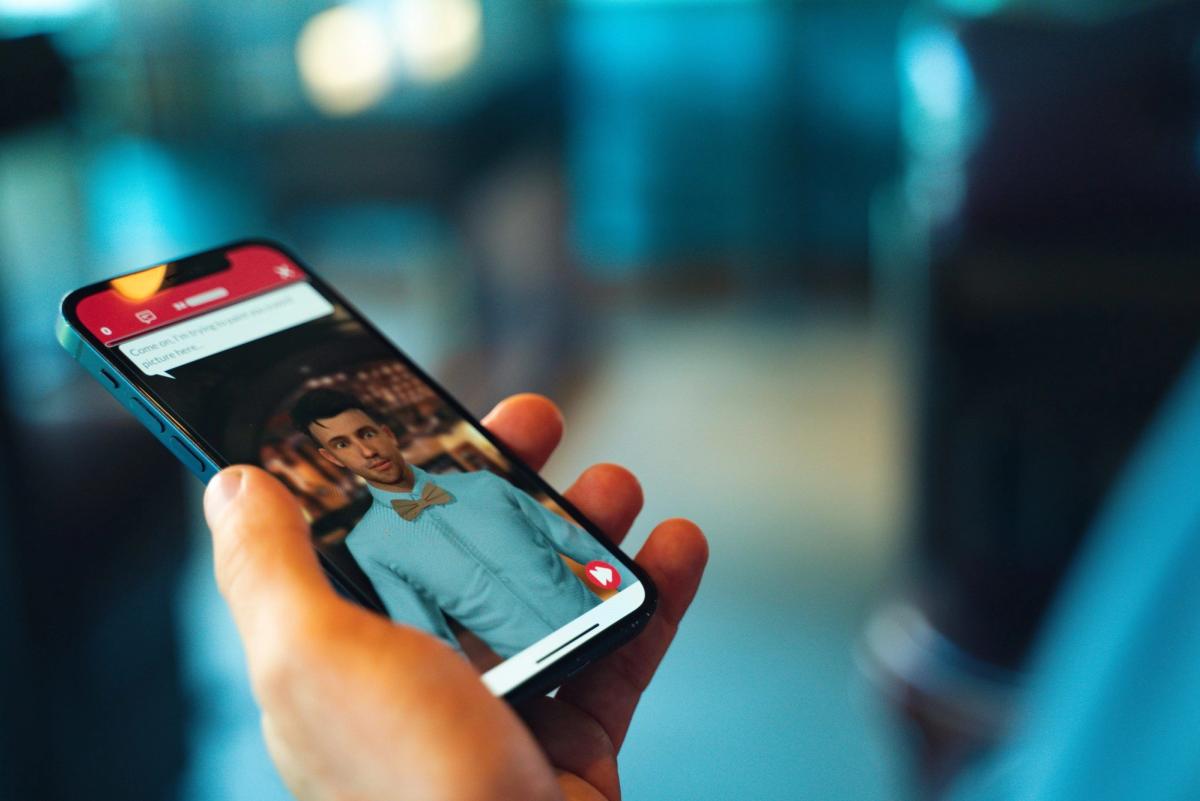 Hand holding a phone showing a 3D character in a light blue shirt and bow tie with a chat message