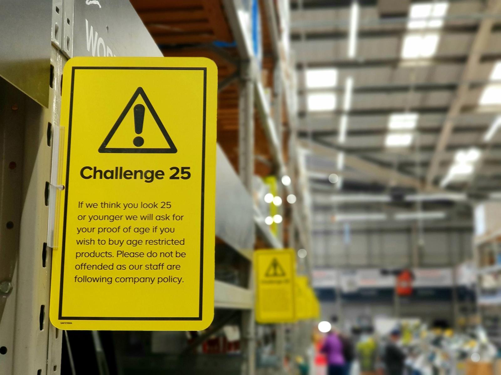 Yellow Challenge 25 age-verification sign on a shelf edge in a store aisle, with a second sign beyond