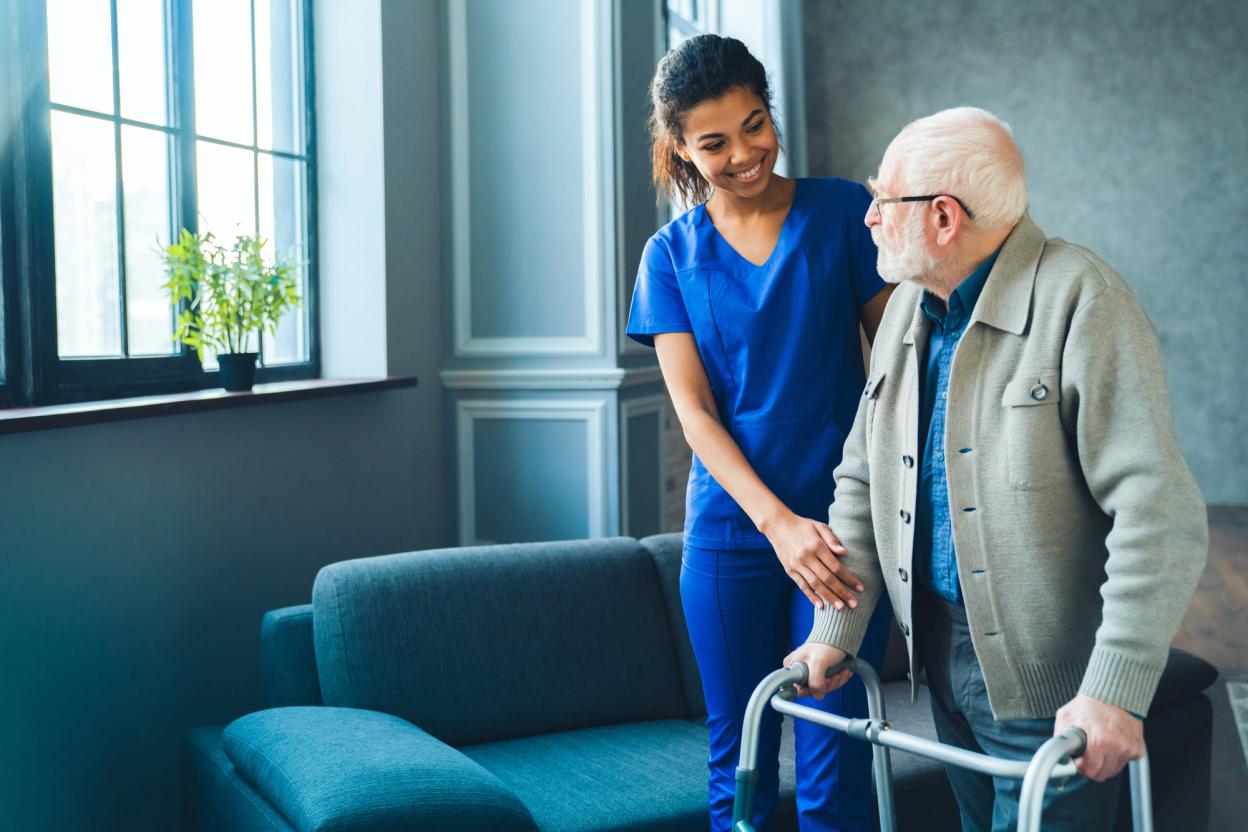 Health worker in blue scrubs supporting an elderly man who is using a walking frame