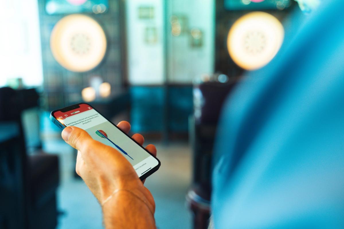Hand holding a phone showing an app screen with a dart illustration, dartboards blurred behind