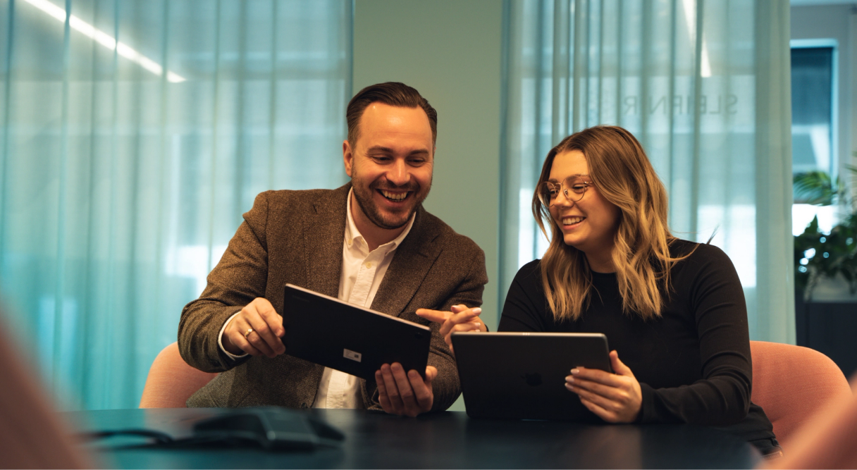 Two colleagues smiling while working together on tablets in a meeting room