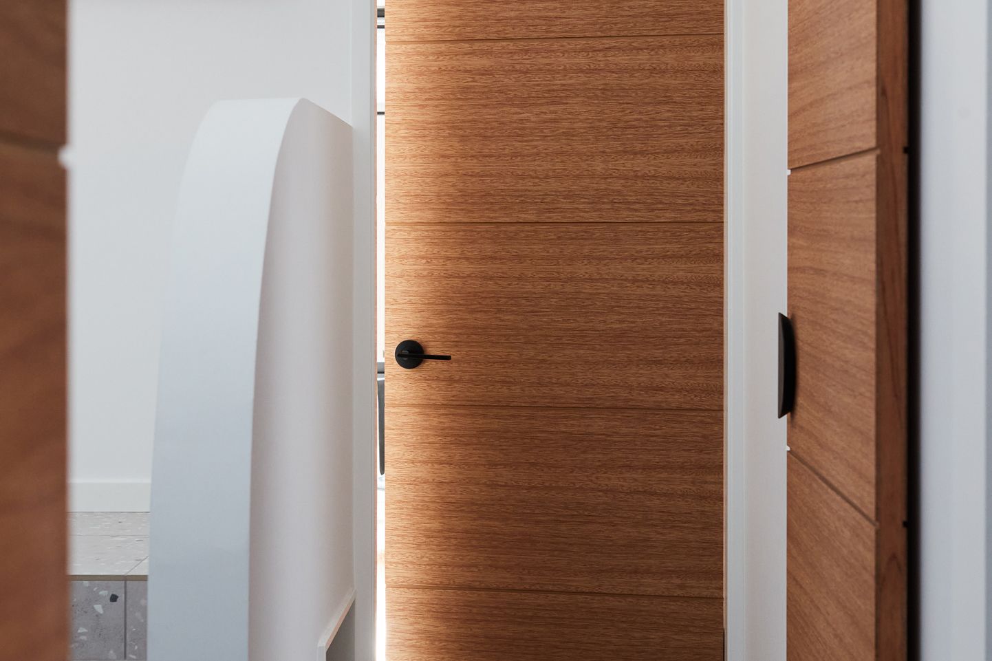 A warm, modern timber interior door HLR250 from the Linear range with horizontal panel detailing and a black lever handle, partially open in a bright hallway. White walls and clean architectural lines create a minimalist, contemporary feel, while the natural wood grain adds warmth and texture. Another matching timber door is visible in the background.