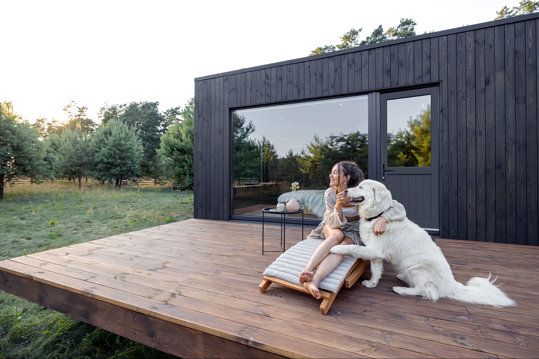 Modern black-clad cabin exterior with a black aluminium VJ panel XE11 door, large floor-to-ceiling window, timber deck, and a woman relaxing outdoors with a large white dog surrounded by natural bushland.