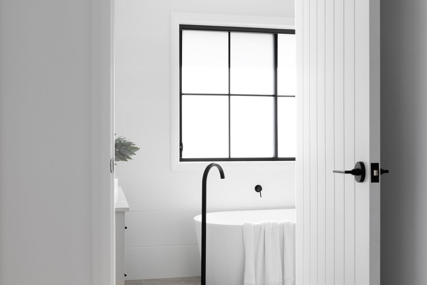 White bathroom with a minimalistic design. Features a black-framed window, a freestanding bathtub with a towel, and sleek black fixtures. Framed by an open HAG11 from the Accent range. Calm and modern ambience.