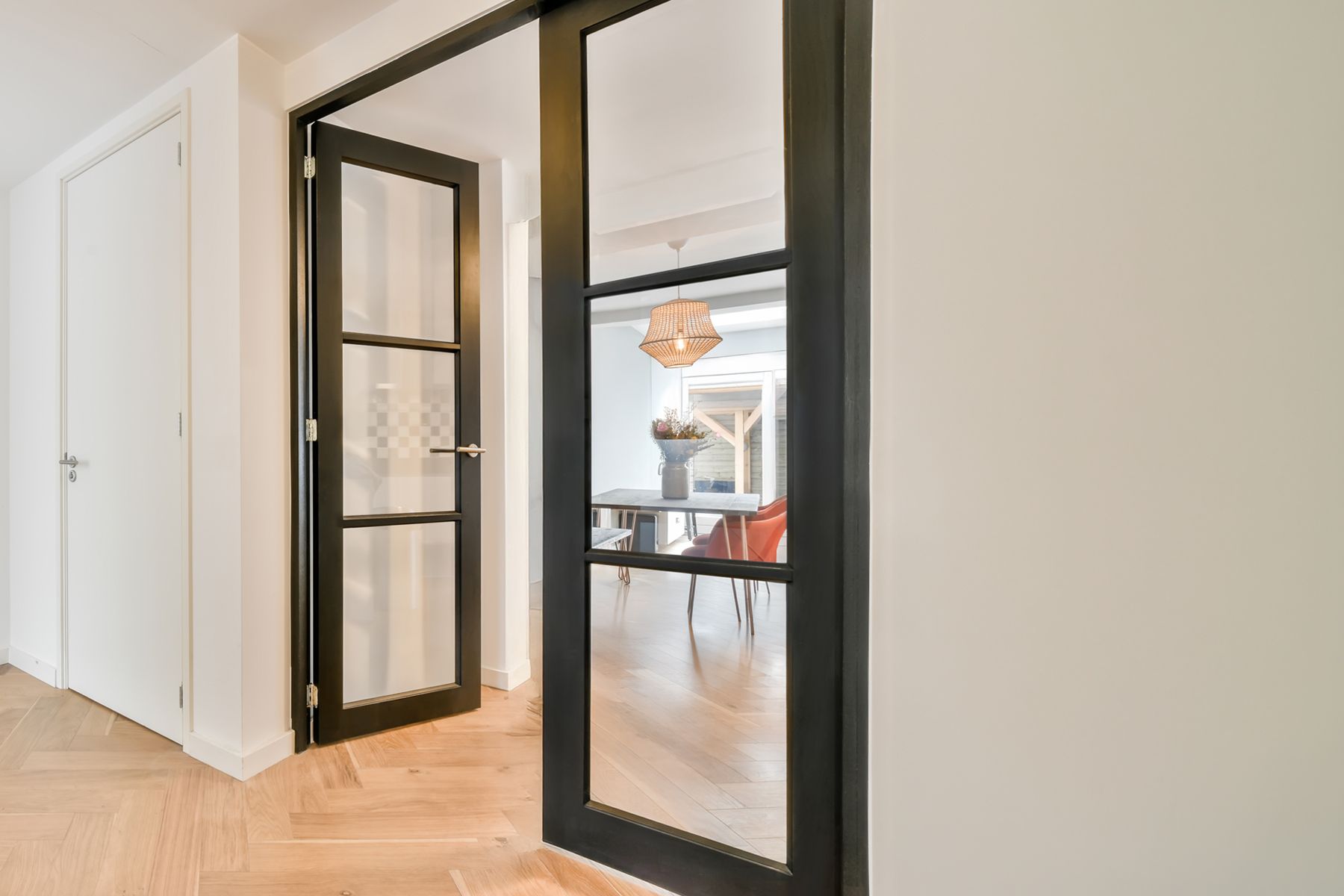 A modern interior with open black framed Allure ALL33 glass doors from the Hume range leading to a bright room. Wooden floors, white walls, and a wicker light fixture creates a cozy atmosphere.