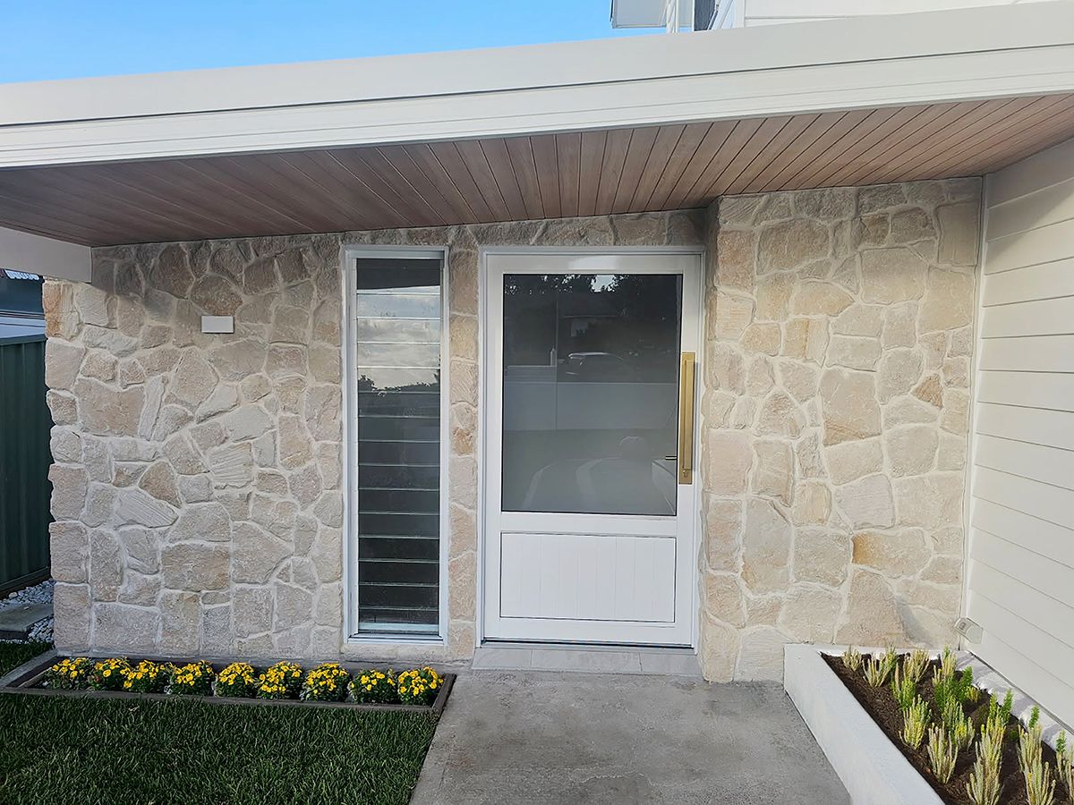 Front entrance of a modern coastal-style home featuring a white aluminium VJ panel XE13 entry door with frosted upper glass, a vertical sidelight, natural stone cladding, timber-look soffit lining, and landscaped garden beds.