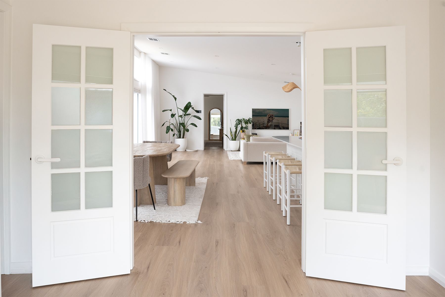 Bayview BAY88 internal double doors in white, featuring French-style panels with translucent glass, opening into a light-filled open-plan living and dining space with warm timber flooring and soft coastal styling.