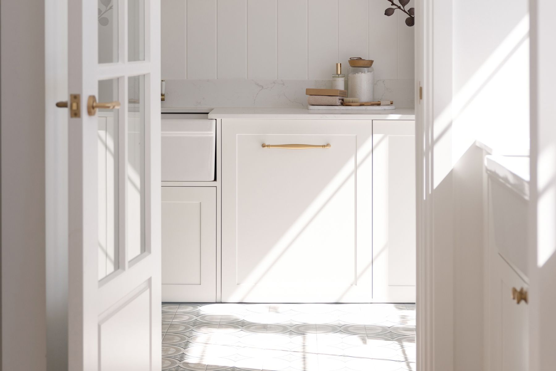 A White Bayview BAY66 door sits ajar. Sunlight streams into a minimalist kitchen with white cabinets, gold handles, and patterned floor tiles. Marble countertop hosts jars and decorative items.