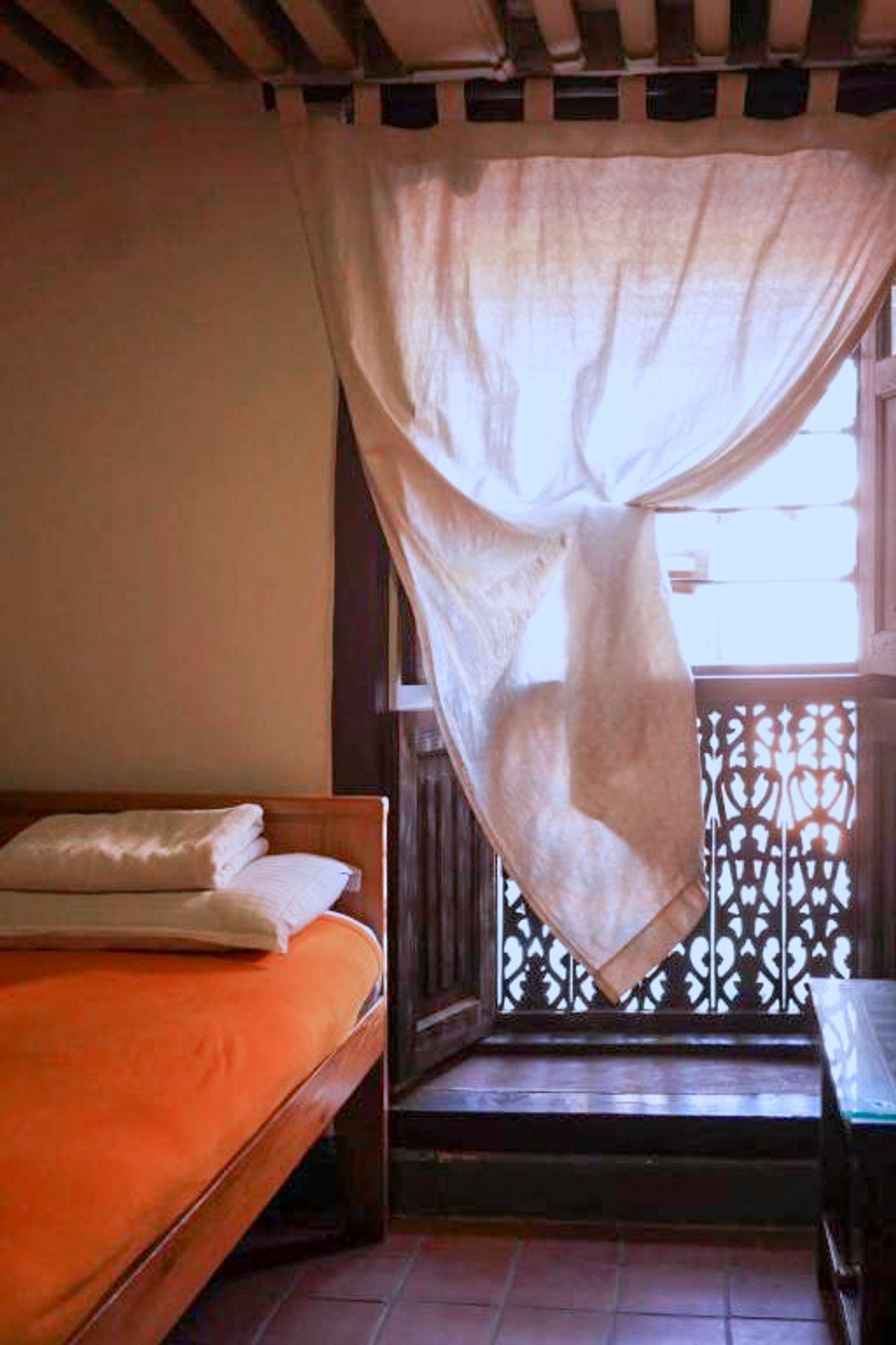 Brahmayani bed beside a carved timber window with a white curtain