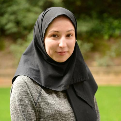 A smiling woman with brown eyes wears a black hijab and a gray sweater, standing in front of a blurred green grass and foliage background.