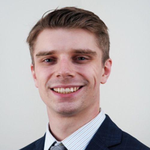 A man with short light brown hair smiles at the camera. He is wearing a dark blue suit, a light blue striped shirt, and a gray tie against a plain light background.