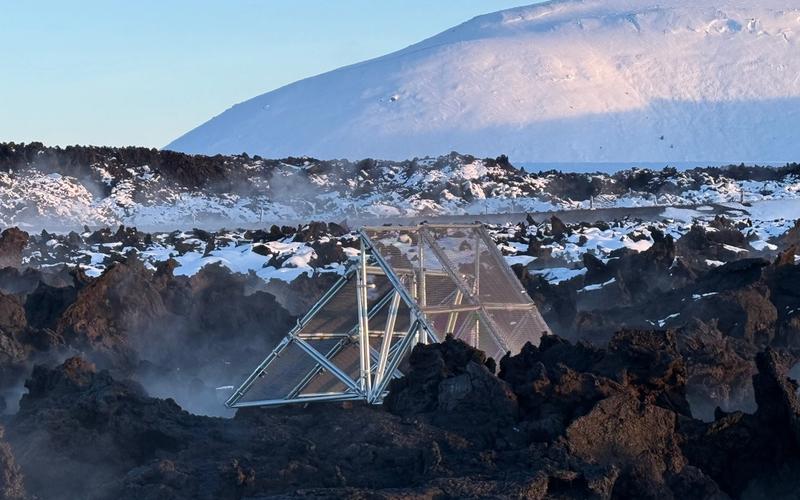 A triangular metal structure partially buried in a rugged volcanic landscape, surrounded by black lava rocks and patches of snow. Steam rises from the ground, and a snow-covered mountain looms in the background under a clear blue sky.
