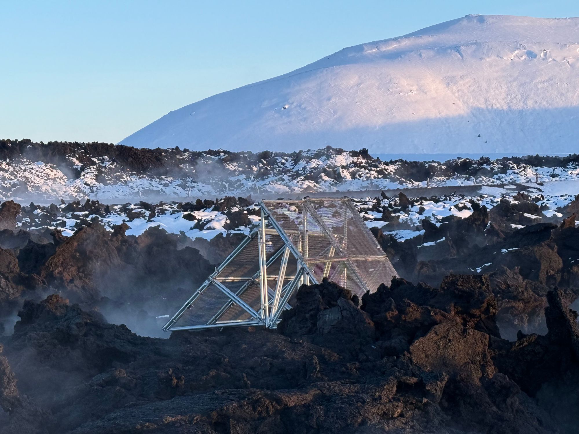 A triangular metal structure partially buried in a rugged volcanic landscape, surrounded by black lava rocks and patches of snow. Steam rises from the ground, and a snow-covered mountain looms in the background under a clear blue sky.