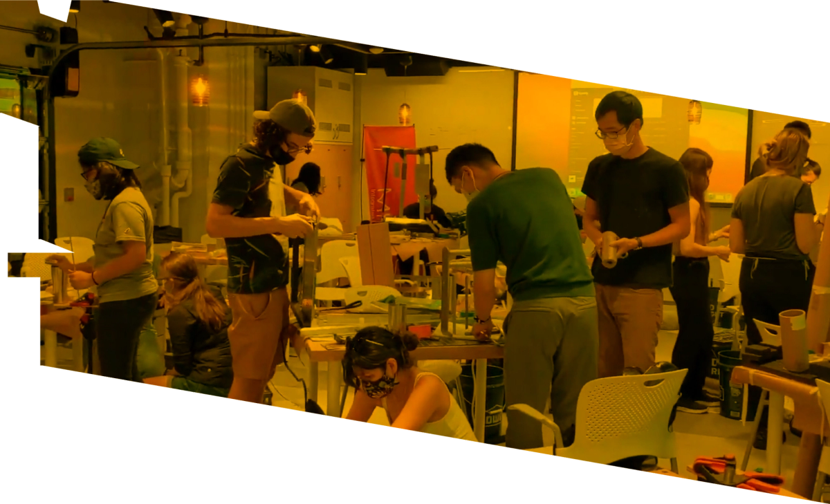 A yellow tinted slightly skewed rectangle image of at least 12 students working in a design lab—constructing architectural models. Each student is wearing a COVID mask, and is working independently.