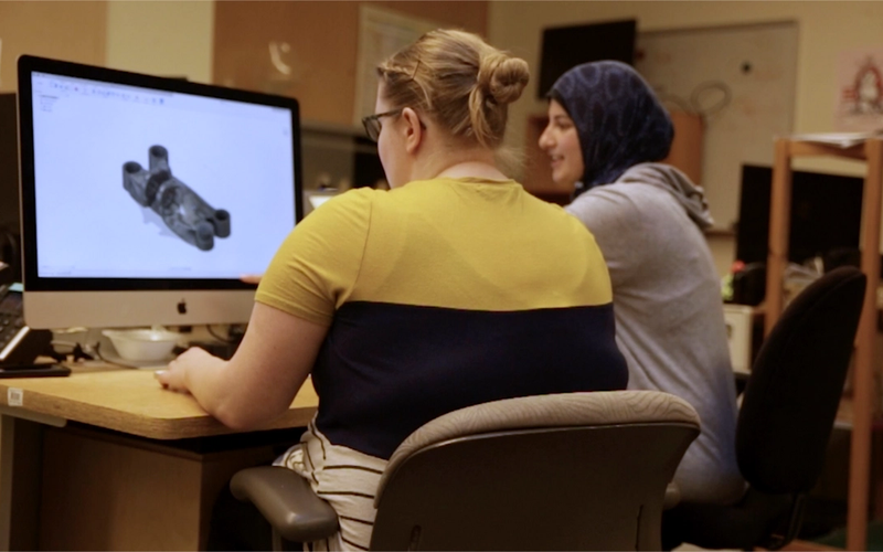 Two women sit at a desk working on a 3D model displayed on a large desktop monitor in a computer lab or design classroom. One is using the mouse to interact with the software, while the other looks on and smiles.
