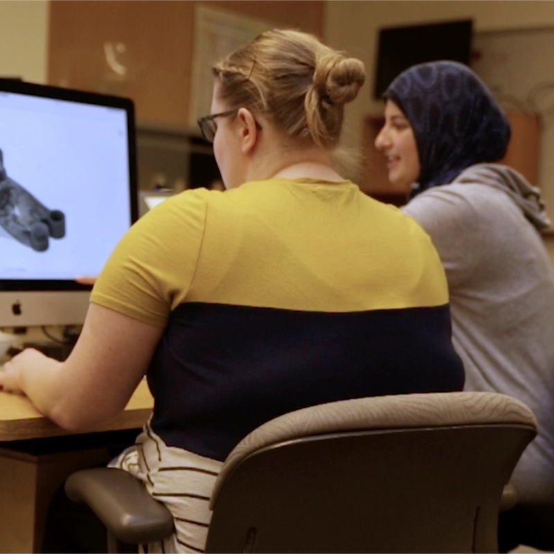 Two women sit at a desk working on a 3D model displayed on a large desktop monitor in a computer lab or design classroom. One is using the mouse to interact with the software, while the other looks on and smiles.