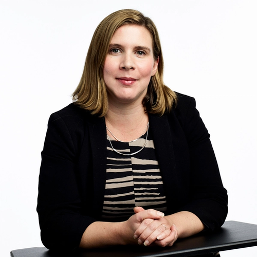 A woman with light brown hair smiles and has her hands clasped on a black surface while wearing a black blazer over a black and tan striped top with a silver necklace, against a plain white background.