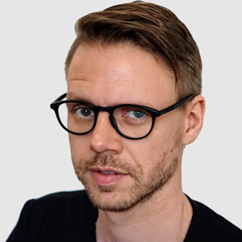 A man with brown hair, a beard, and glasses, wearing a black shirt, looks toward the camera in a close-up headshot against a plain gray background.