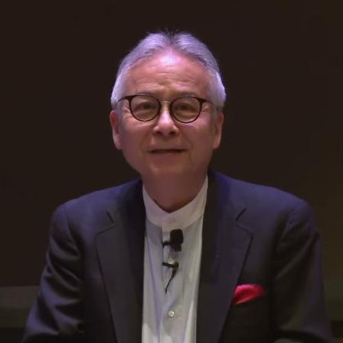 A smiling man with gray hair and glasses wears a black suit jacket over a light blue shirt with a red pocket square, sitting in front of a dark brown background with an MIT sign visible on the right.