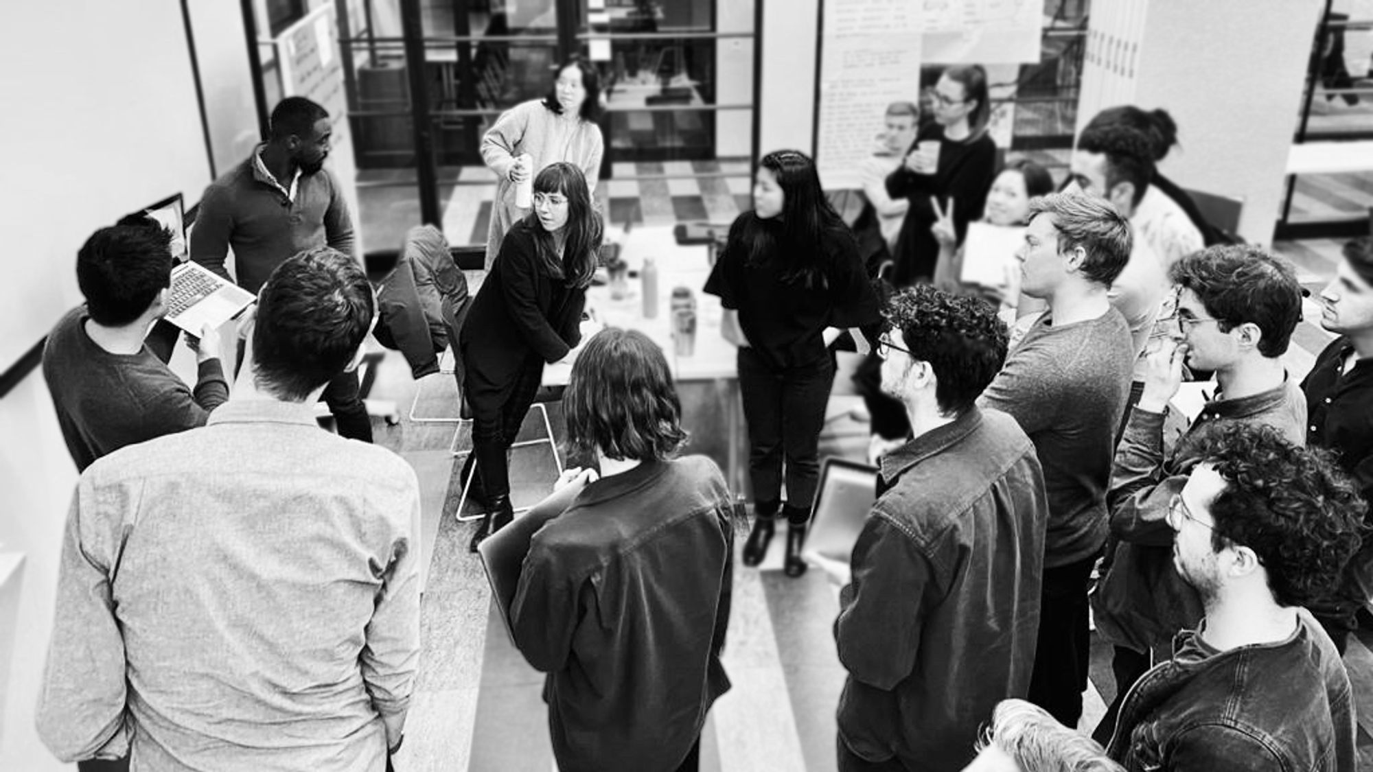 A group of about fifteen people stand in a semicircle around a presenter during a workshop or discussion. The presenter gestures toward a whiteboard while others listen, some holding laptops or notebooks. The setting appears to be a collaborative classroom or studio with glass walls and posters in the background.