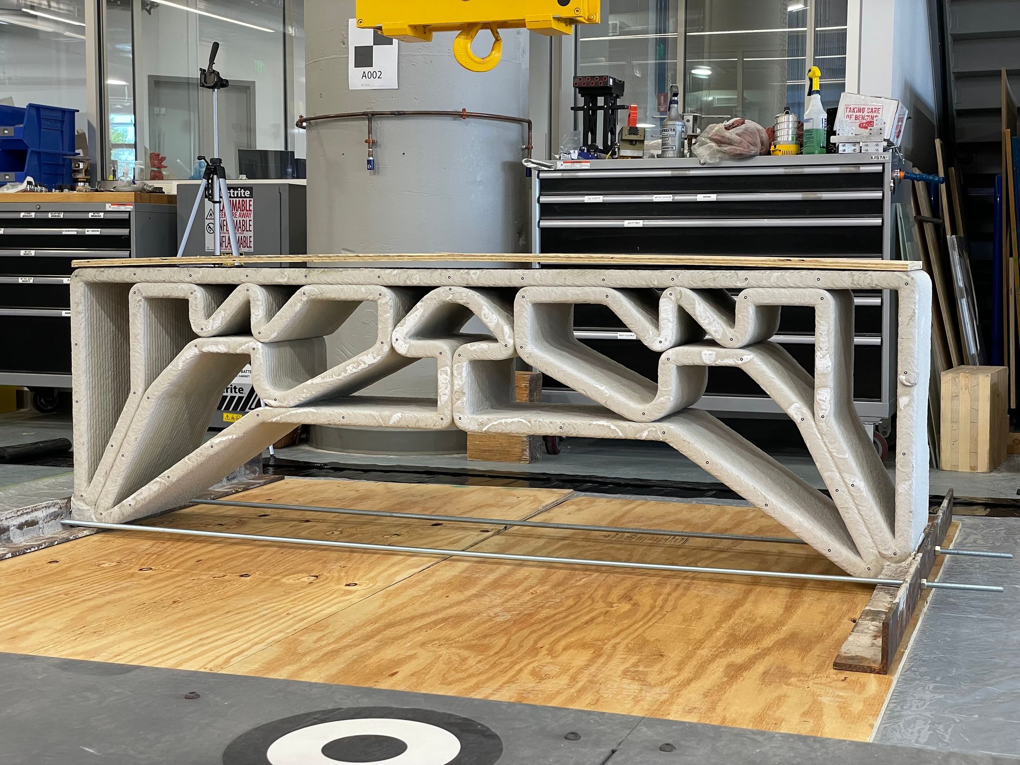 3D-printed concrete beam with intricate lattice-like internal supports, resting on a wooden platform in a workshop setting, surrounded by tools and equipment.