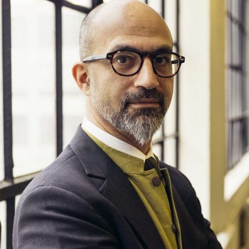 A bald man with a graying beard and glasses, wearing a dark gray jacket over a gold-colored vest and white shirt, looks seriously at the camera in front of a large window with gridded panes.