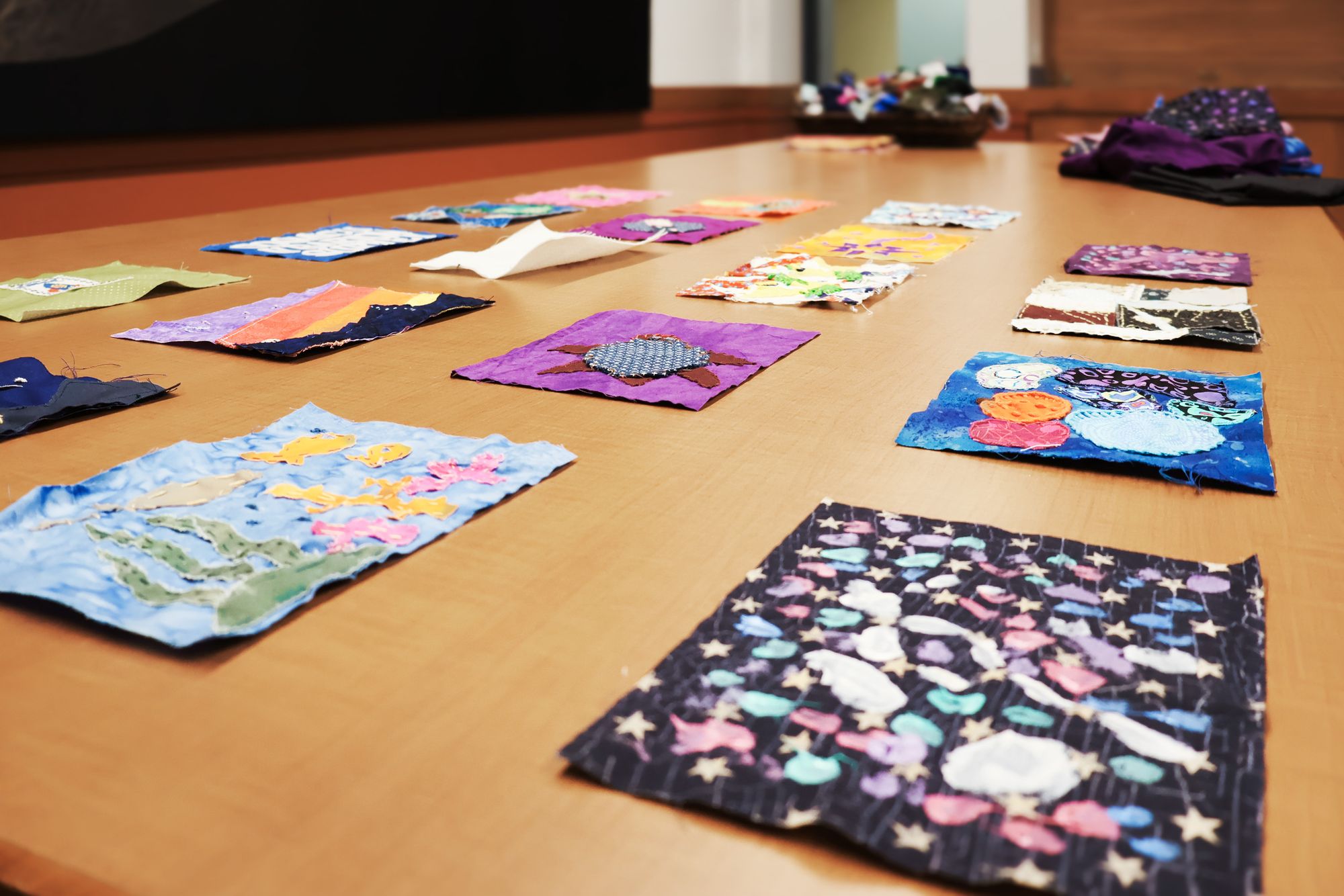 Colorful quilt squares are laid out across a wooden table, each decorated with unique stitched patterns, abstract shapes, and nature-inspired motifs.