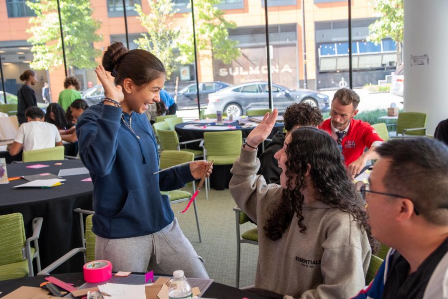 A group of students share a laugh while working on a hands-on design activity. One student raises her hand for a high-five, while another meets it enthusiastically. Colorful materials such as tape and paper are spread across the tables, and other groups can be seen collaborating in the background of the bright, glass-walled space.