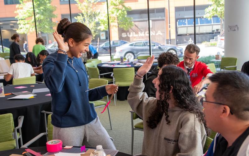 A group of students share a laugh while working on a hands-on design activity. One student raises her hand for a high-five, while another meets it enthusiastically. Colorful materials such as tape and paper are spread across the tables, and other groups can be seen collaborating in the background of the bright, glass-walled space.