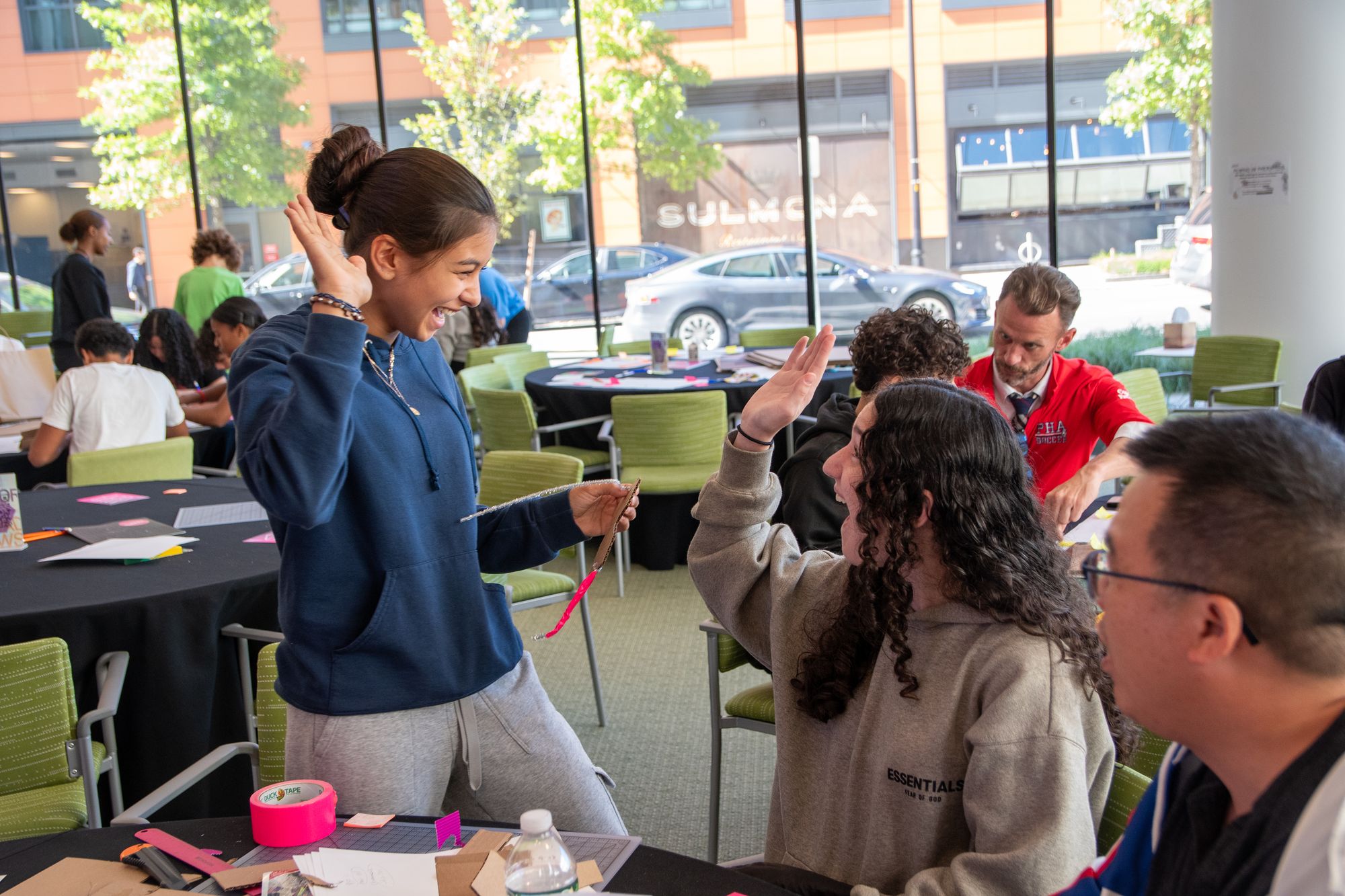 Two teenage girls giving each other a high five during a workshop session in a room with green chairs and tall glass windows. 