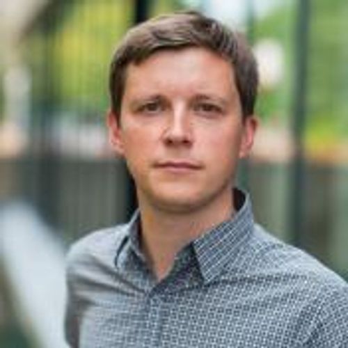 A man with brown hair and a serious expression, wearing a patterned gray button-down shirt, poses for a headshot outdoors with a blurred background of green foliage and a building.
