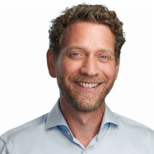 A man with blue eyes, a beard, and curly brown hair, smiles widely at the camera while wearing a light blue button-down shirt against a plain white background.