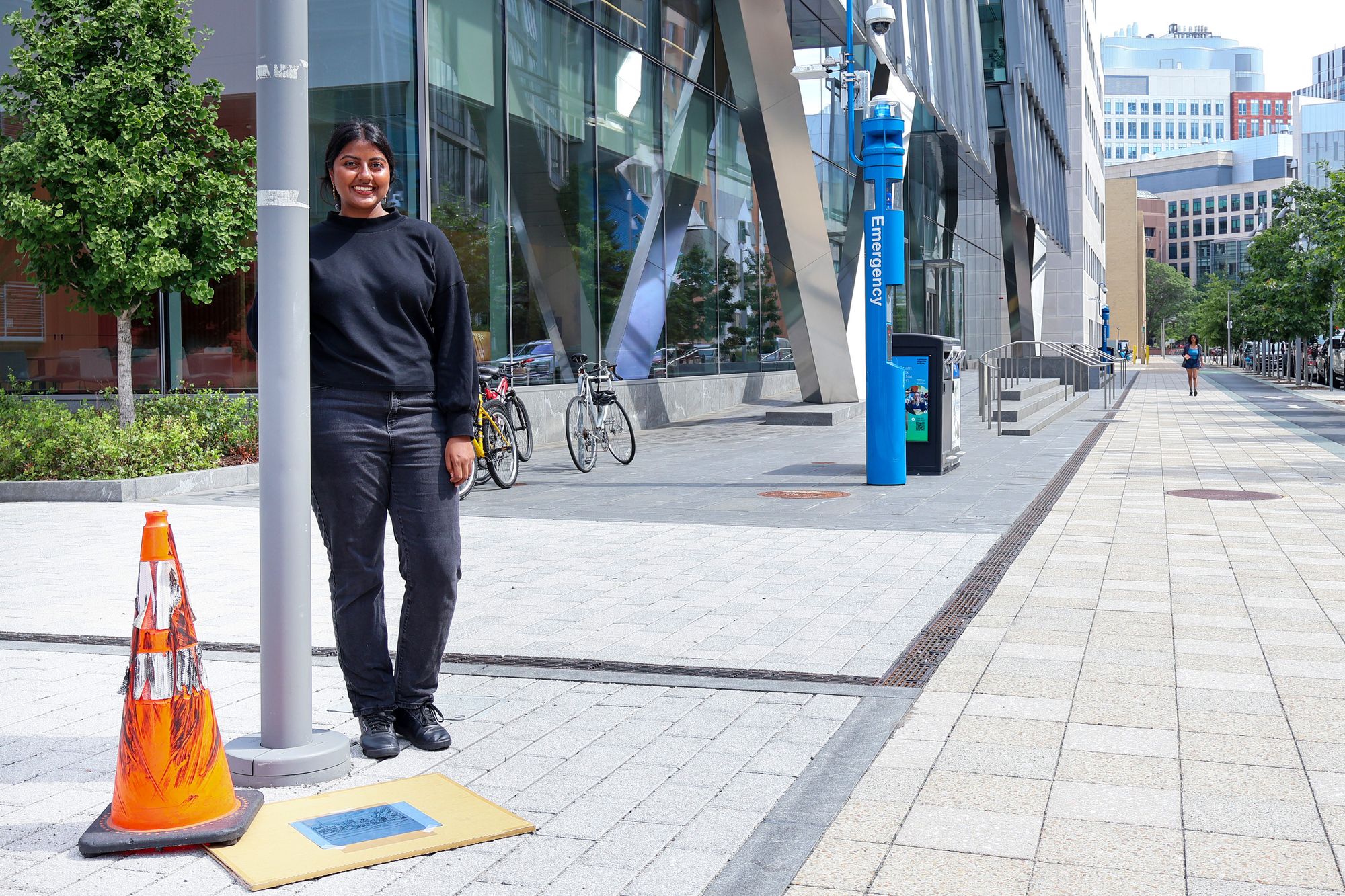 A woman stands outdoors on a city sidewalk, leaning against a lamppost with an orange traffic cone and a drawing taped on the ground. A modern glass building and bicycles are behind her.
