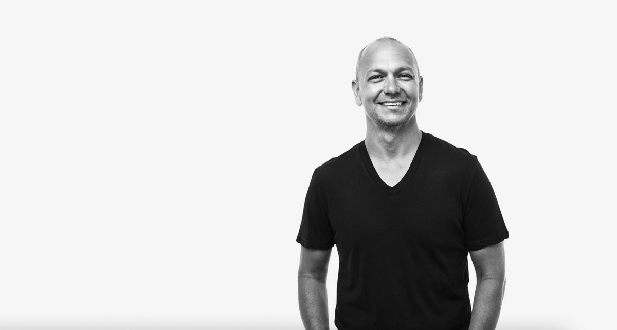 Black-and-white portrait of Tony Fadell, smiling and standing casually with one hand in his pocket, wearing a plain black V-neck T-shirt against a light background.