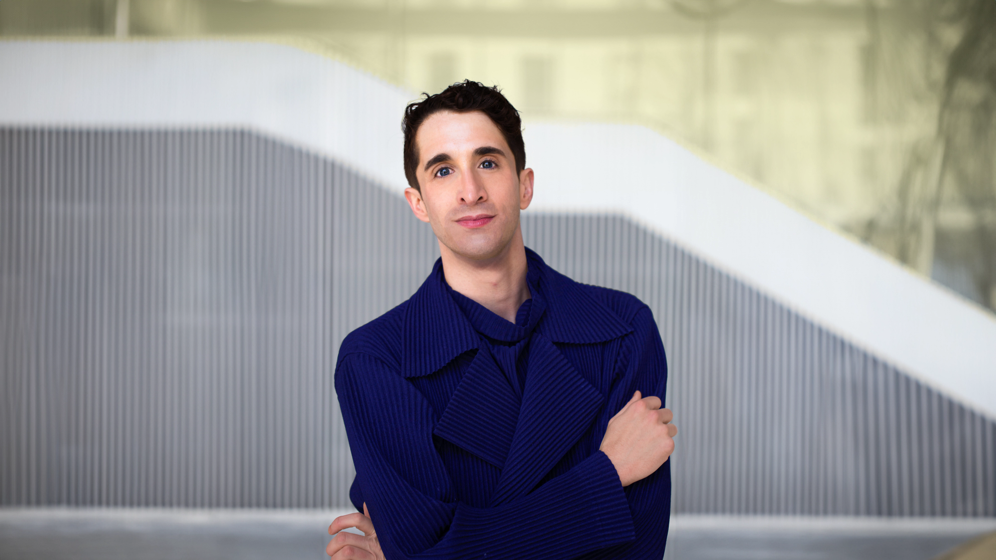 Person standing in front of a modern architectural background with diagonal white and gray striped elements. They are wearing a deep blue, textured coat with a layered collar, and are looking at the camera with a composed expression, one arm crossed over their body.