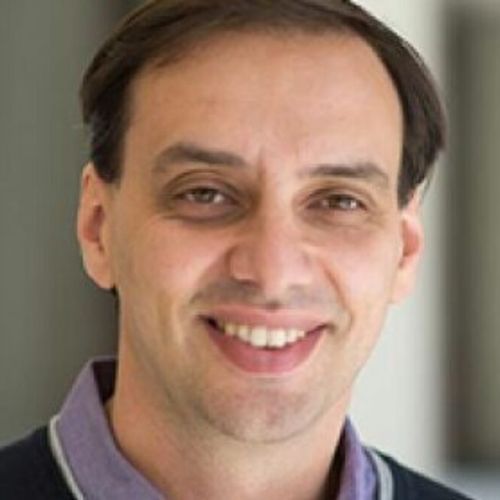 A man with dark hair and a wide smile wears a dark sweater over a light purple shirt, posing for a headshot with a blurred light-colored background.