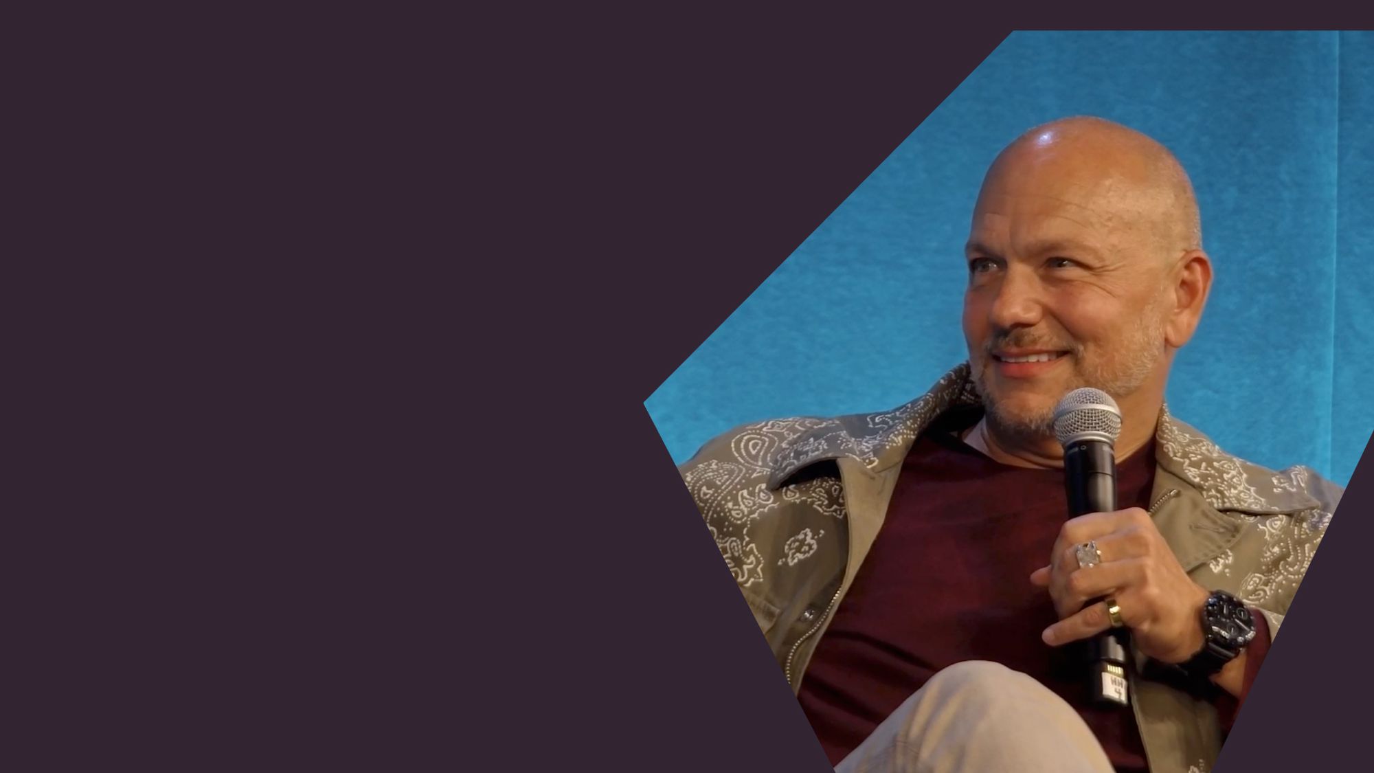 A photo of Tony Fadell sitting and holding a microphone, against a blue background