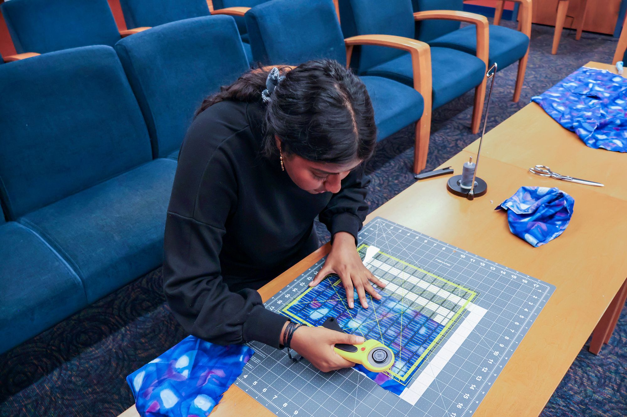 A woman in a black top leans over a table, using a rotary cutter and ruler to cut blue patterned fabric on a gridded cutting mat.