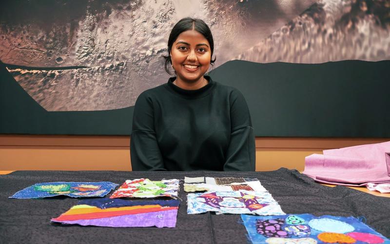 A young woman with dark hair tied back, wearing a black top, sits smiling at a table covered with colorful fabric swatches. The fabrics feature bright patterns in blue, purple, red, and multicolored designs. Behind her is a large wall image with a textured, abstract surface in shades of gray and black.