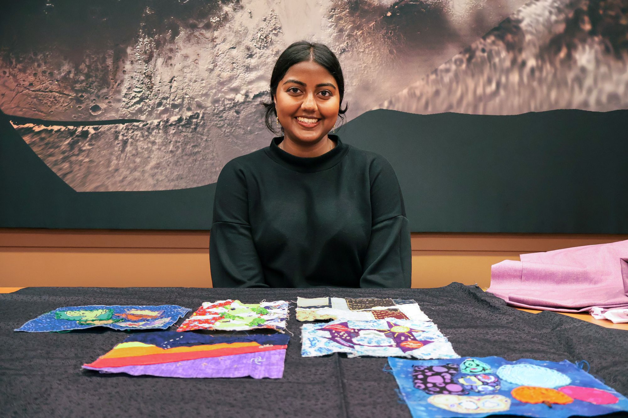 A young woman with dark hair tied back, wearing a black top, sits smiling at a table covered with colorful fabric swatches. The fabrics feature bright patterns in blue, purple, red, and multicolored designs. Behind her is a large wall image with a textured, abstract surface in shades of gray and black.