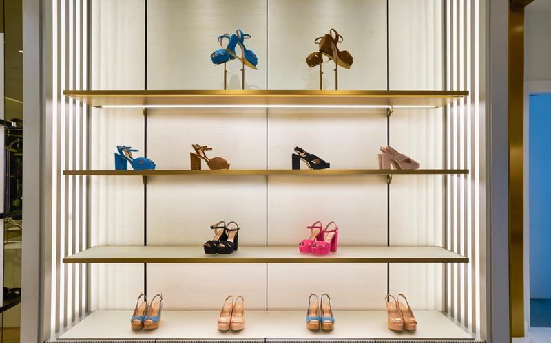 Display of Stuart Weitzman platform sandals in various colors, including blue, black, brown, pink, and nude, arranged neatly on illuminated gold-framed shelves inside a boutique.