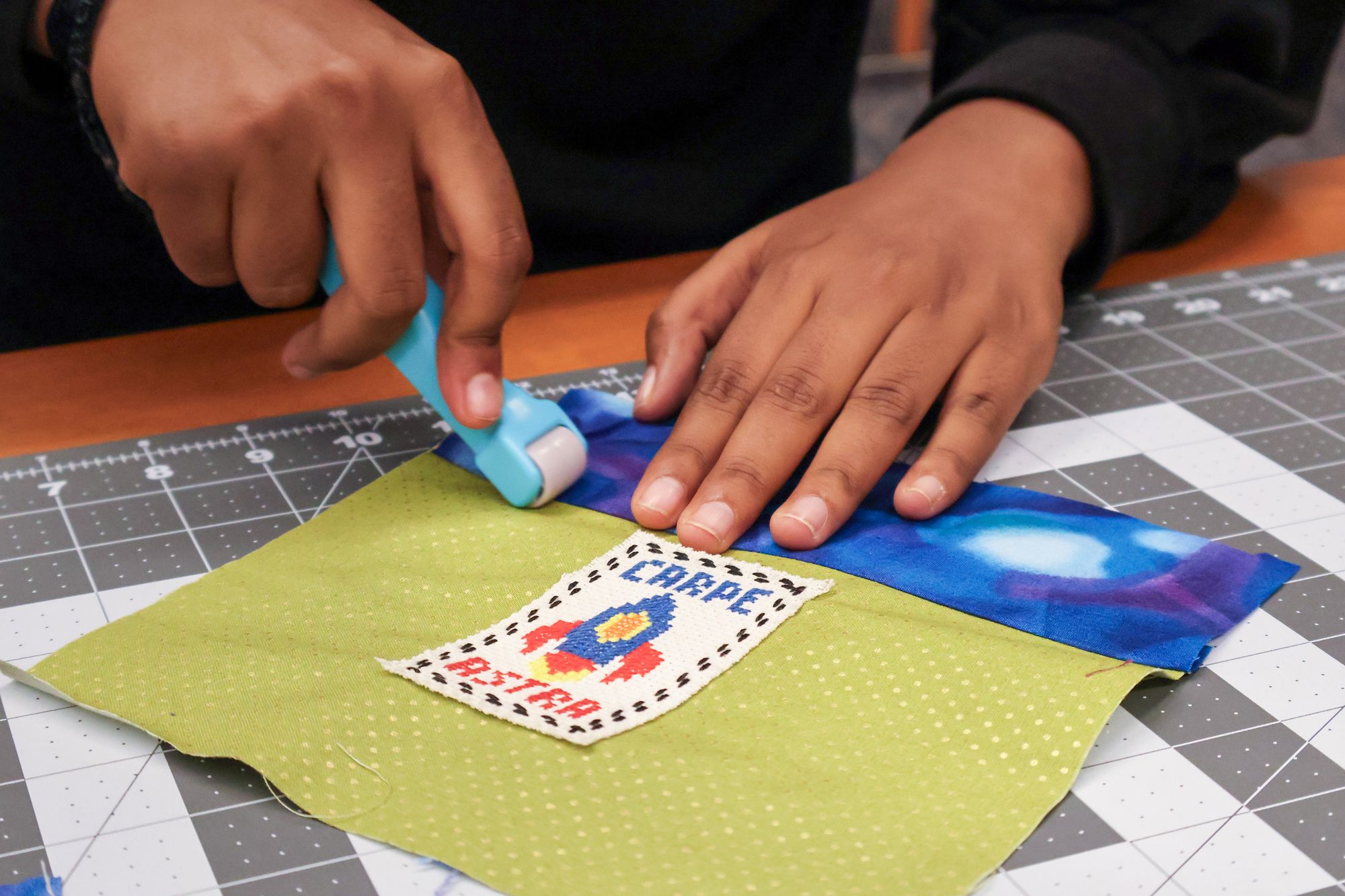 Hands pressing fabric with a seam roller on a green piece of cloth stitched with a rocket patch that reads “CARPE ASTRA.”