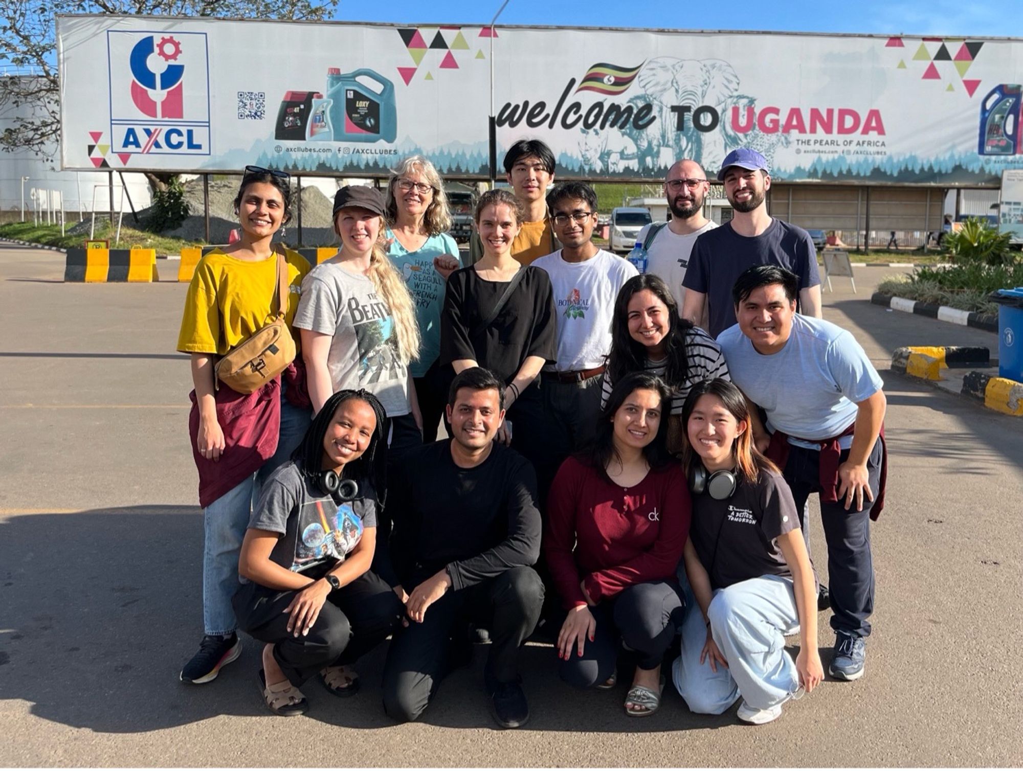A diverse group of fifteen people poses for a group photo in front of a large sign reading “Welcome to Uganda – The Pearl of Africa.” They are smiling and appear to be part of a team or program visiting the country, with clear skies and an industrial backdrop behind them.
