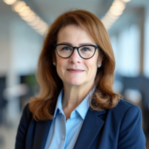 A woman with auburn hair and glasses smiles at the camera, wearing a navy blue blazer over a light blue collared shirt, with a blurred indoor office background.