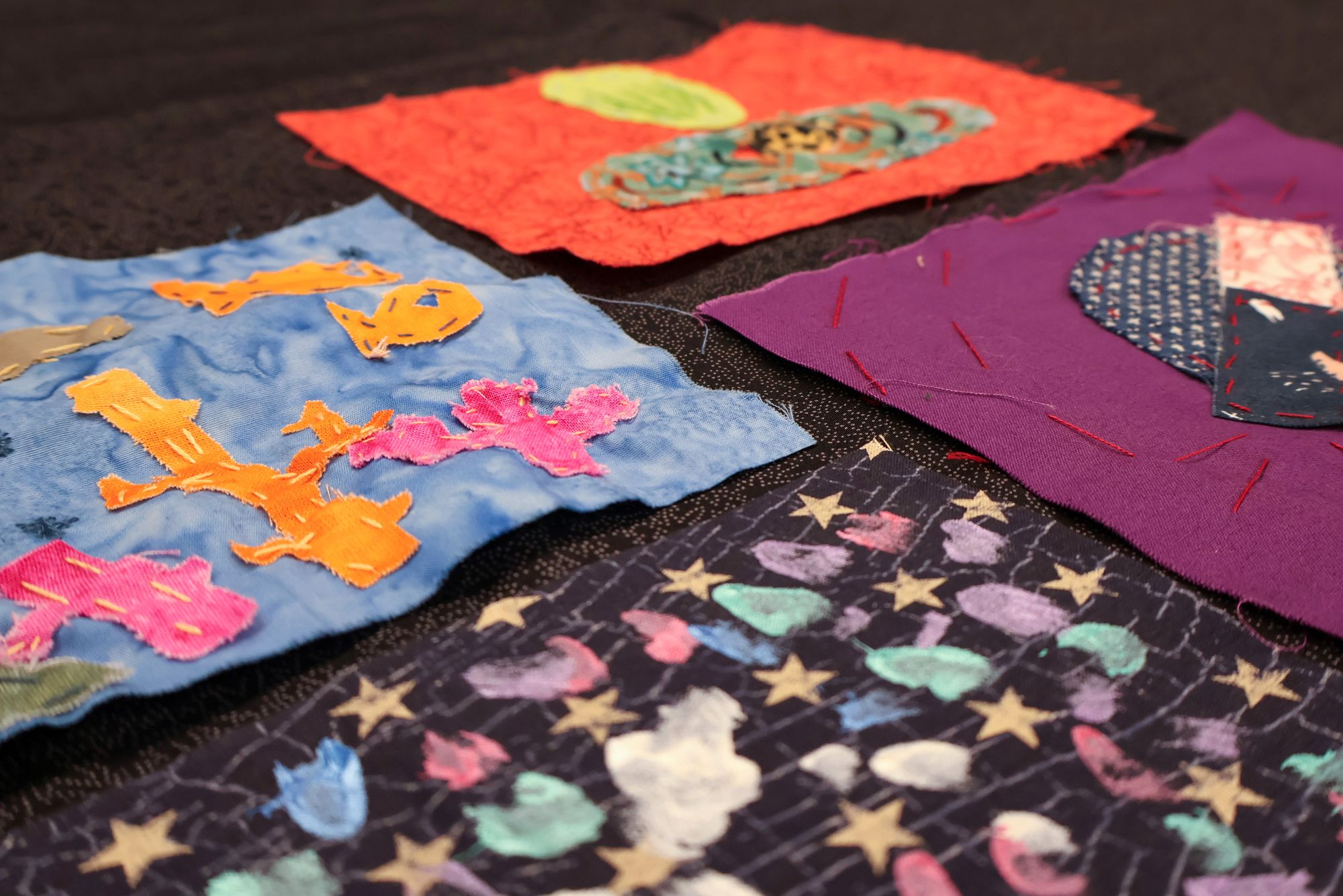 A group of colorful quilt squares laid out on a dark surface, featuring stitched fish, planets, abstract shapes, and patterns.