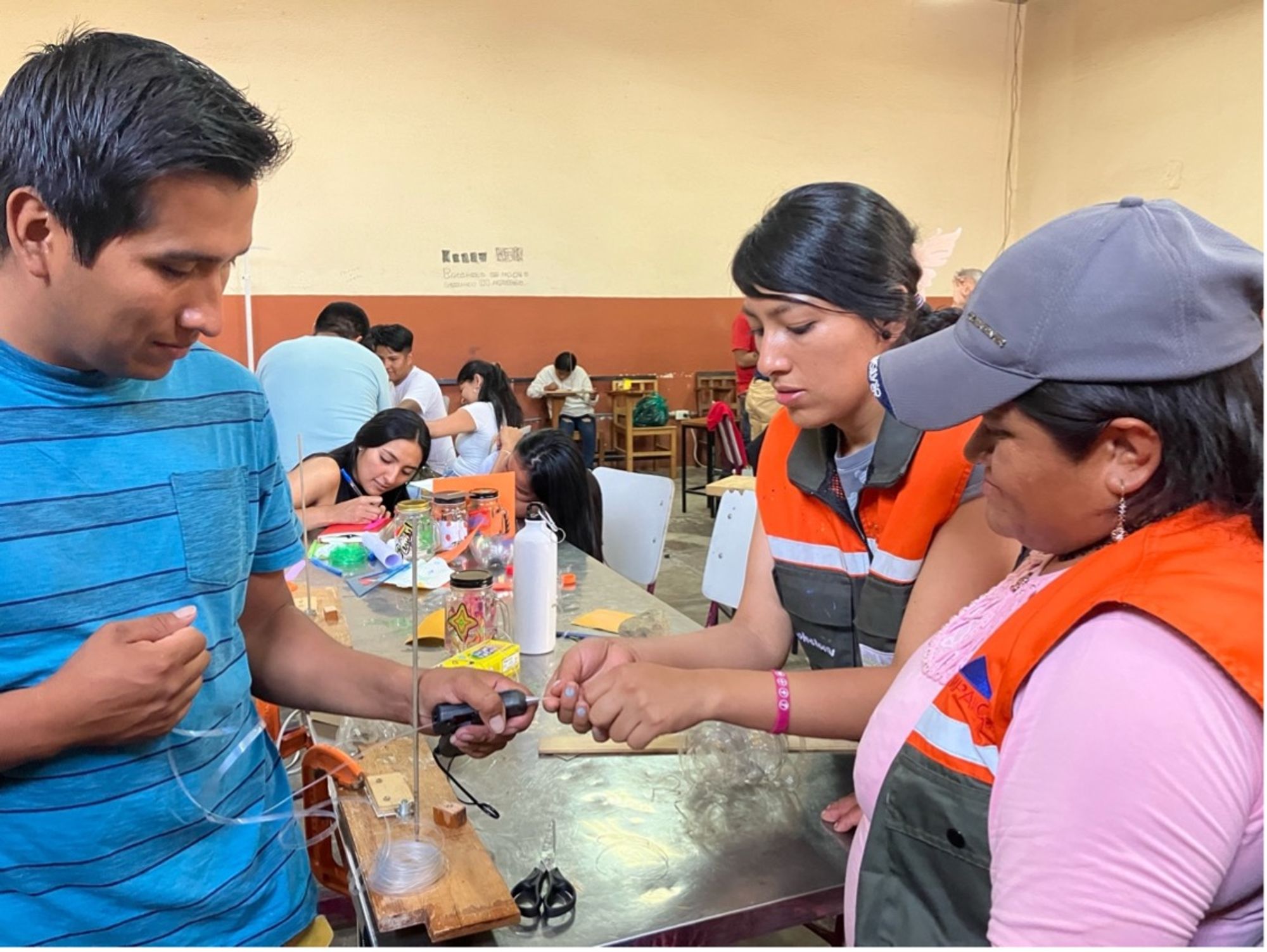 Three people, two wearing orange safety vests, collaborate around a table while assembling or testing a small DIY science or engineering device involving tubing and a plastic container. Other participants are visible in the background working on projects in a classroom setting.