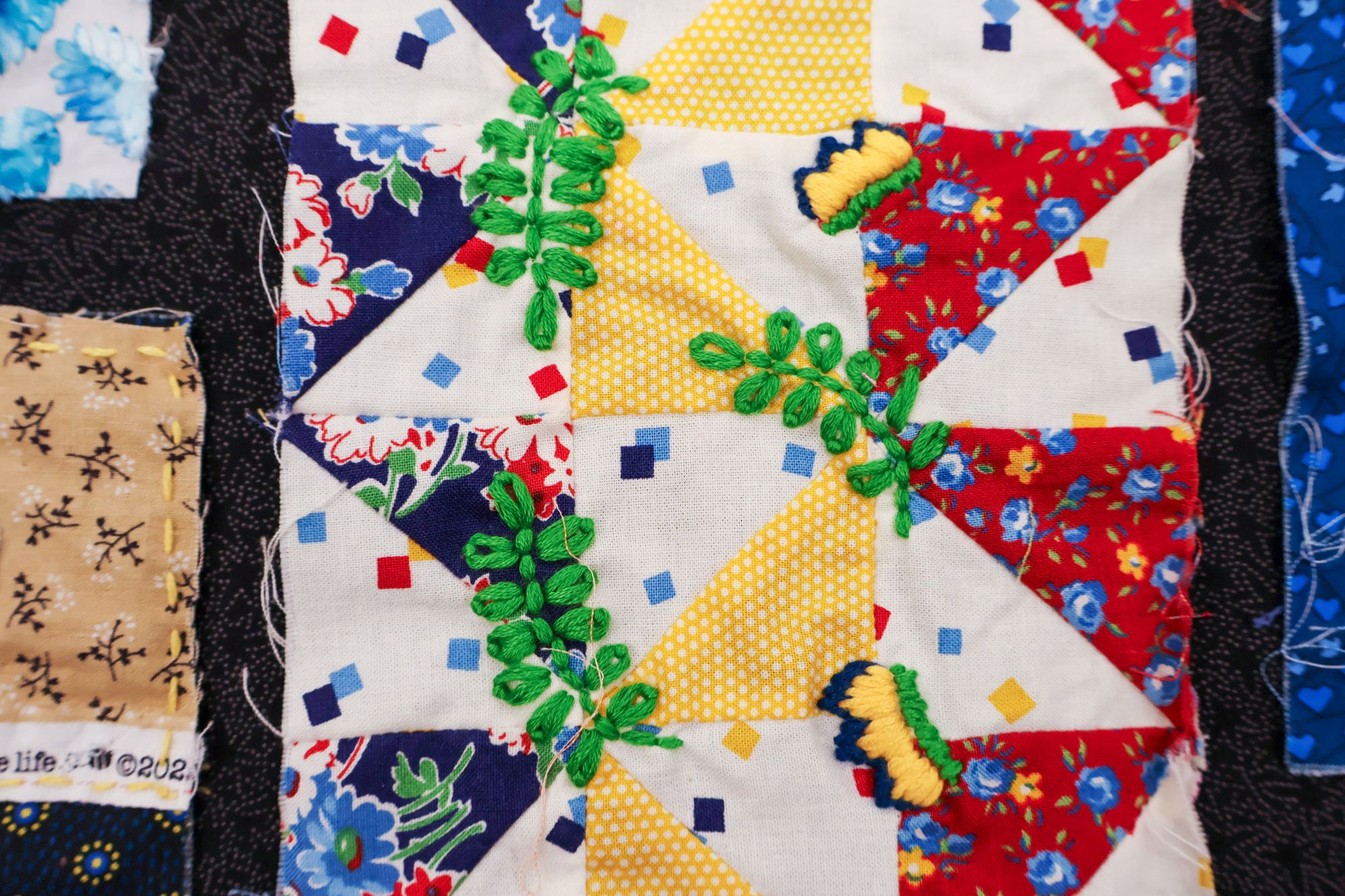 A close-up of a quilt square decorated with red, yellow, and white patterned triangles, overlaid with green embroidered leaves and a small butterfly.